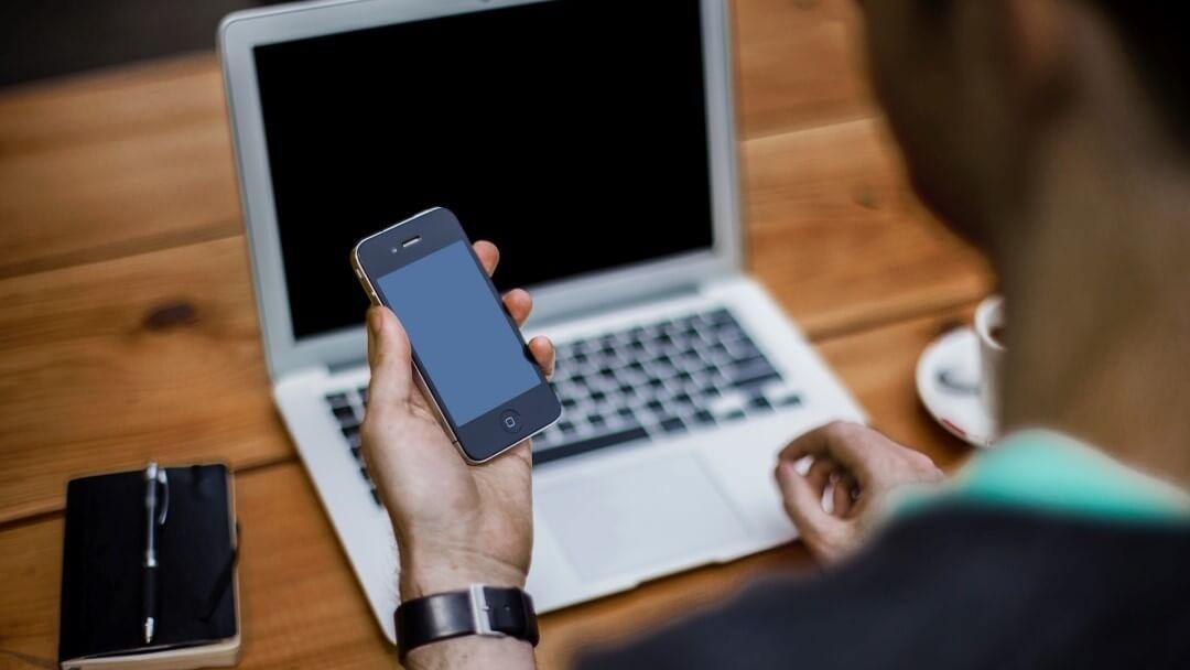 Image symbolique: un homme est assis devant un ordinateur portable et regarde son smartphone qu'il tient à la main. Les deux écrans sont noirs.