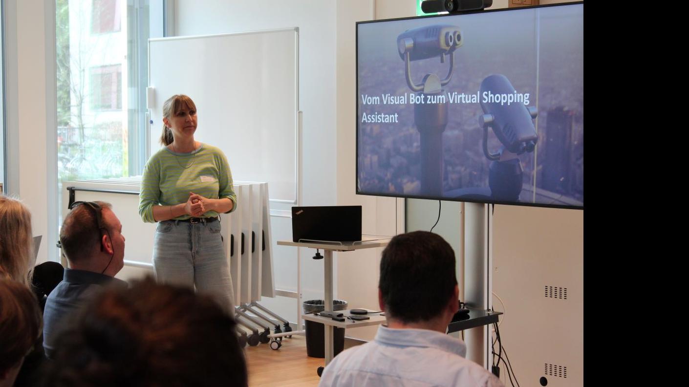 Une femme présente le projet informatique au public, dont quatre personnes sont visibles. L'écran affiche "Du Visual Bot à l'assistant virtuel d'achat".