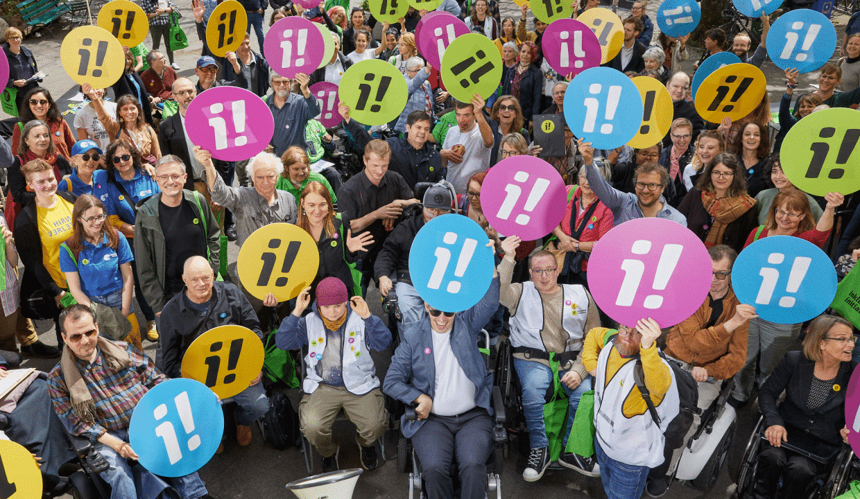 Menschenmenge mit bunten Schildern in der Hand, auf denen das Logo der Inklusions-Initiative zu sehen ist (zwei Ausrufezeichen, eins davon steht auf dem Kopf)