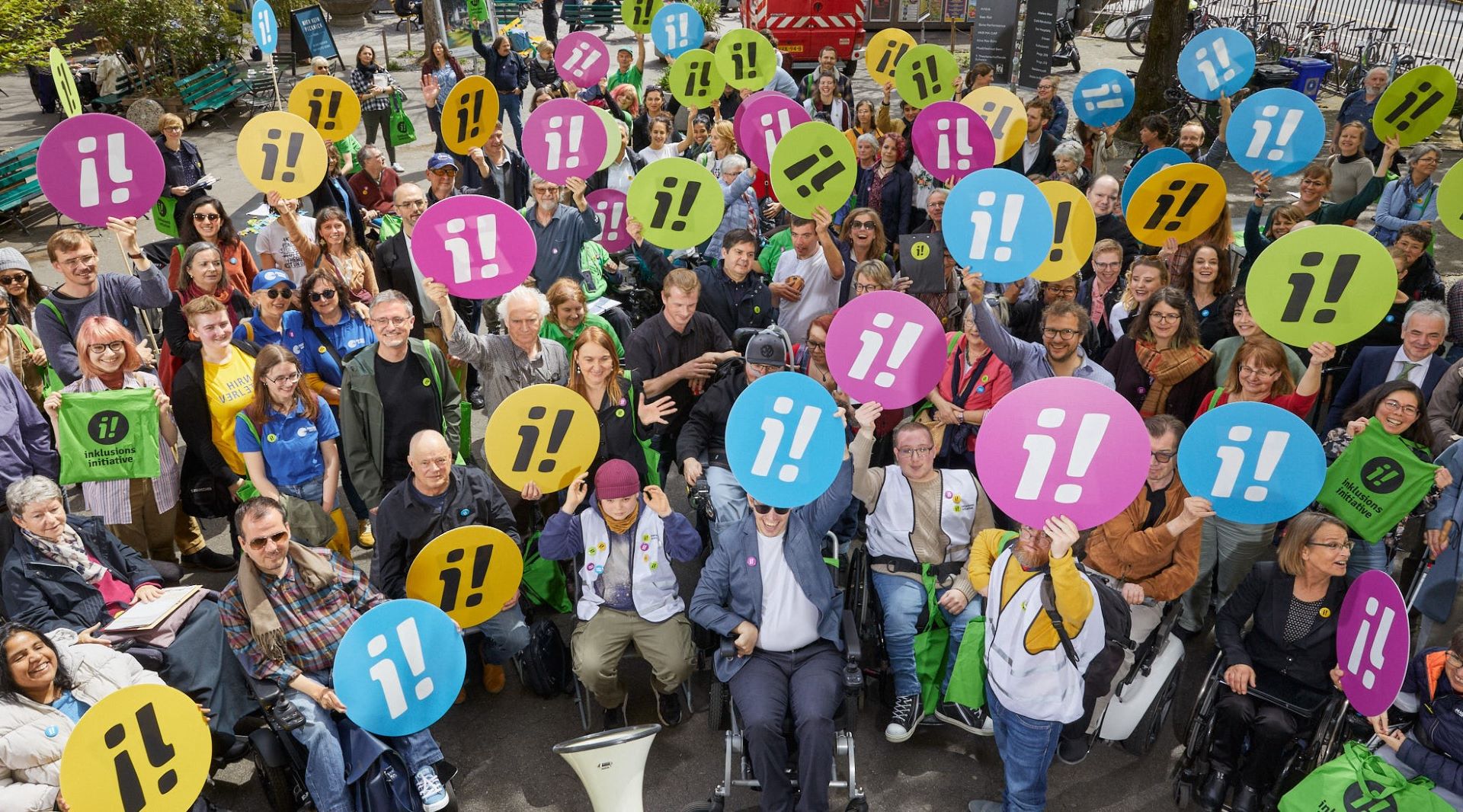 Rund 100 Personen mit verschiedenen Behinderungen halten verschiedenfarbige, runde Schilder mit dem Logo der Inklusions-Initiative 