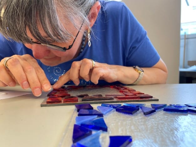 Workshops im BBZ Zürich. Eine stark sehbehinderte Frau mit blauem Hemd beugt sich nah an das von ihr gefertigte Kunstwerk. Vorsichtig legt sie rote und blaue Glasscherben auf eine transparente Glasplatte, um ein Glasfusing-Produkt herzustellen.