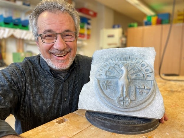 Kunst im BBZ Zürich: Ein etwa 60-jähriger Mann mit Brille strahlt stolz in die Kamera. Vor ihm auf der Werkbank präsentiert er sein während einem Jahr angefertigte kurdische Kunstfigur - ein Pfau mit aufgeschlagenem Schwanzfedern und historischen Symbolen.