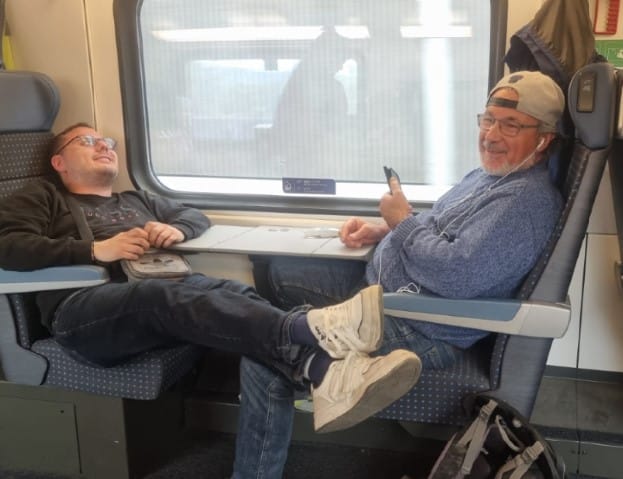 Das BBZ auf Reisen. Ein jüngerer und ein älterer Mann - beide sehbeeinträchtigt - sitzen sich im Zugabteil gegenüber. Der jüngere scheint friedlich zu schlafen, der ältere ist mit seinem Handy beschäftigt.