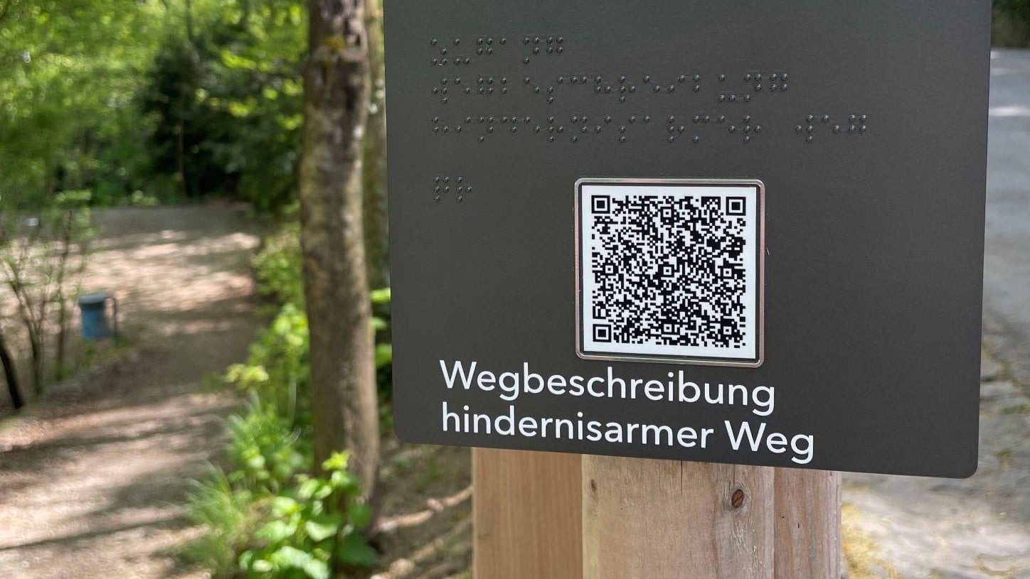 Der Ausschnitt einer dunkelfarbigen Tafel beim Eingang der Englischen Anlage. Darauf steht in Brailleschrift der Text: hindernisarmer Weg. rechts davon befindet sich der QR-Code welcher mit einem taktil erfassbaren Rahmen umrandet ist. Darunter steht der Text in Schwarzschrift. Links von der Tafel sieht man den Weg in den Garten.