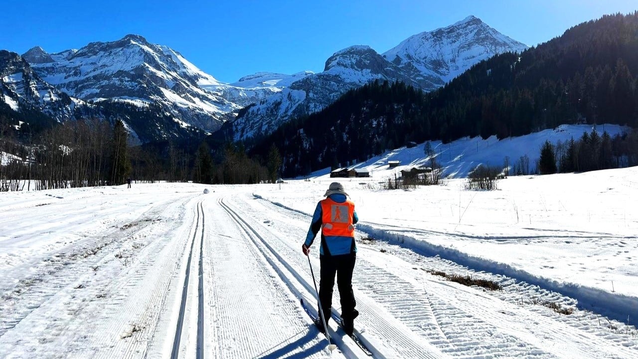 Eine Person bewegt sich mit Langlaufschiern auf einer Loipe in einer winterlichen Landschaft vorwärts. Die Person trägt eine orange Warnweste mit dem Piktogramm einer Person mit weissem Stock darauf.