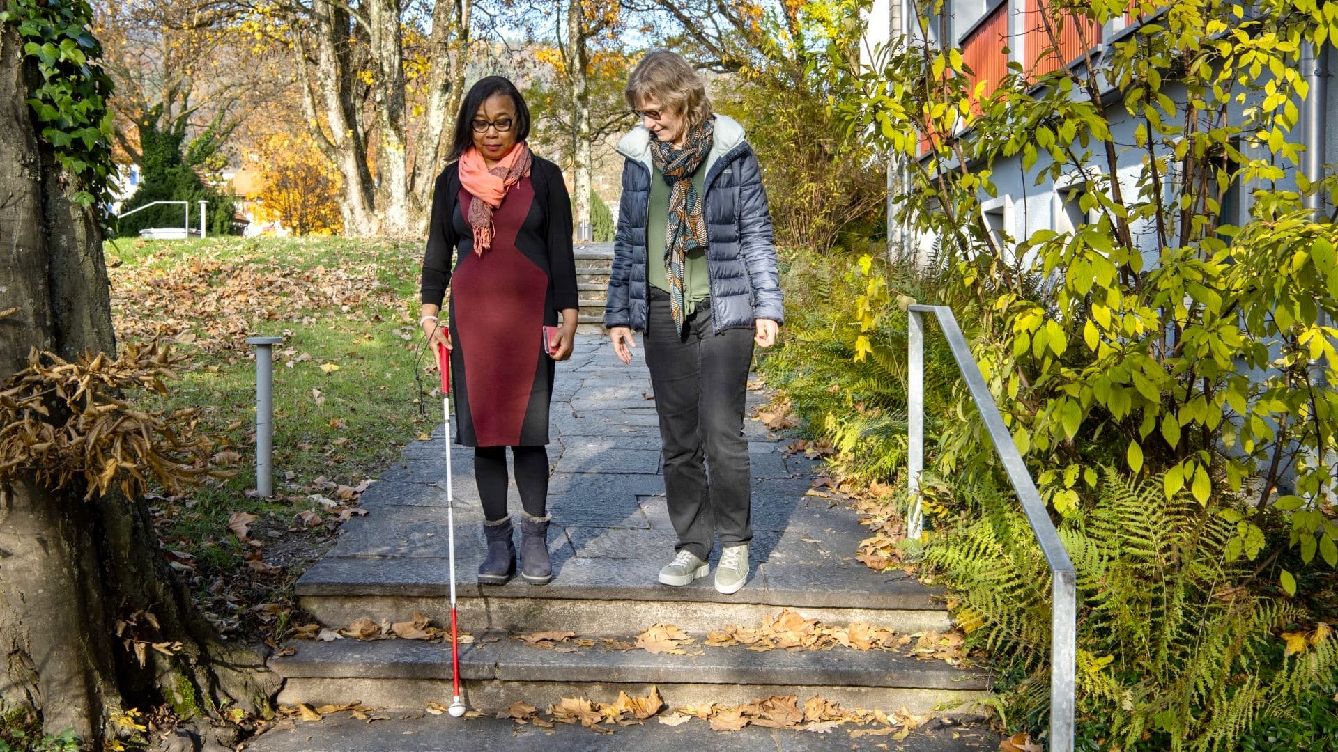 Eine Frau mit einer Seheinschränkung läuft mit einem weissen Stock gemeinsam mit einer Instruktorin eine Treppe hinunter.