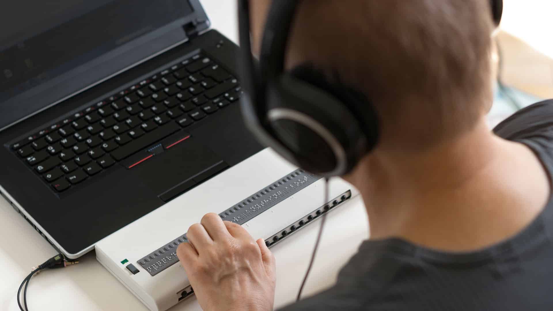 Eine Person arbeitet mit Kopfhörern und einer Braillezeile an einem Laptop.