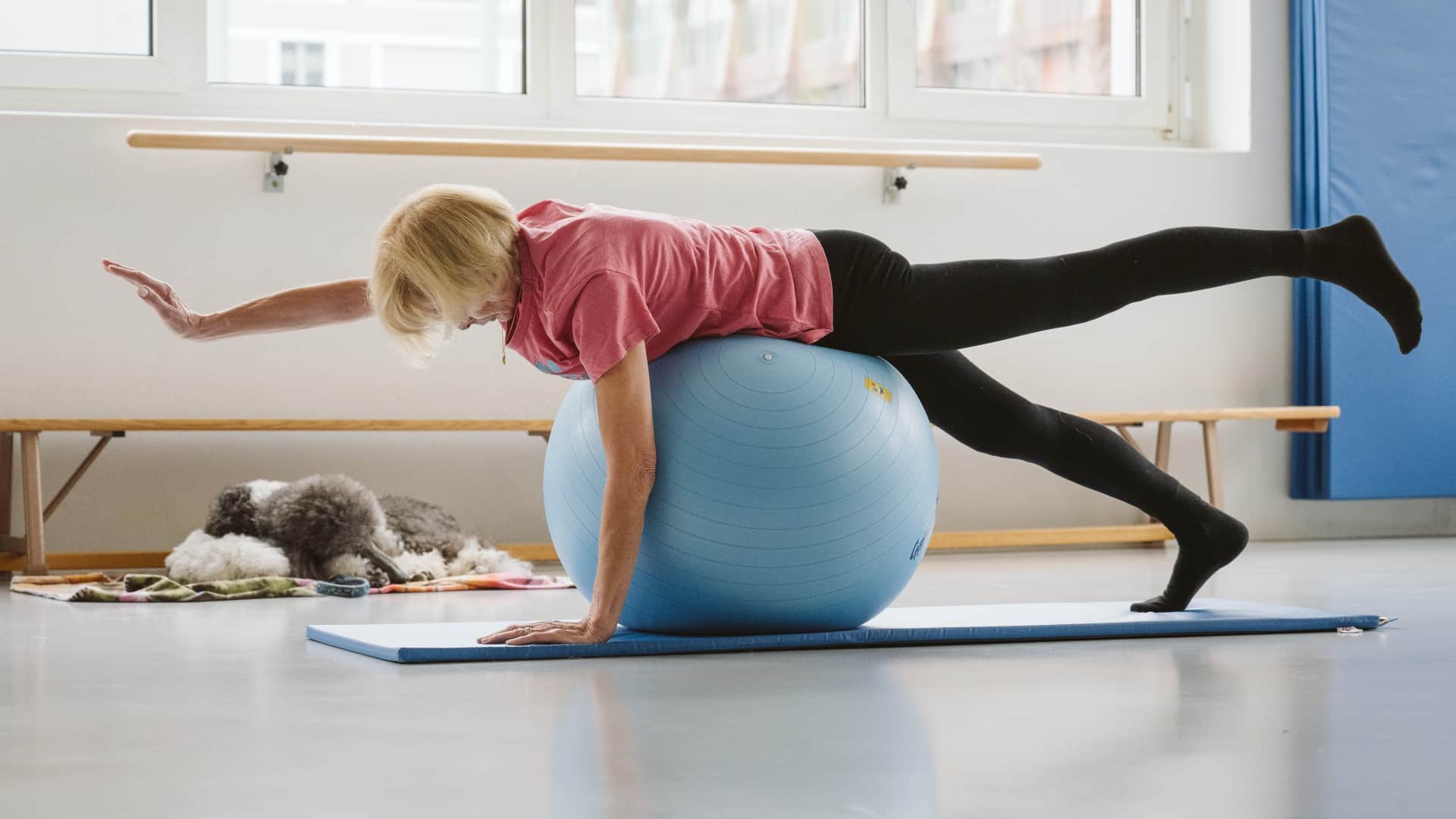 Trainingsraum: Eine ältere Frau in Trainings-Kleidung liegt mit dem Bauch nach unten quer auf einem hellblauen Gymnastikball und macht Gymnastikübungen. Im Hintergrund liegt ein Blindenführhund auf dem Boden.