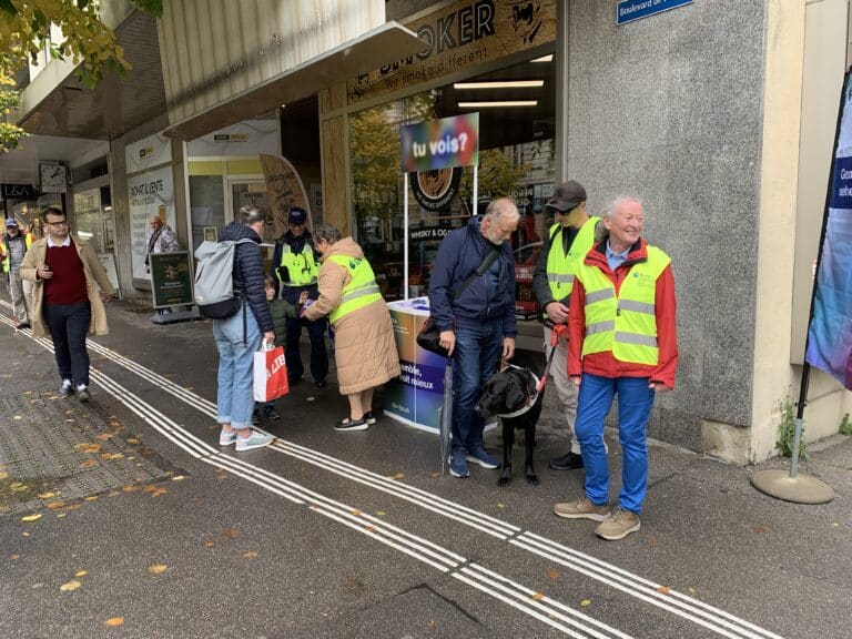 Dieses Bild zeigt eine Gruppe von Personen, von denen einige gelbe Westen tragen, in der Nähe eines Informationsstands auf einer Straße, mit einem Blindenführhund und einem Schild im Hintergrund, auf dem "tu vois?" steht.