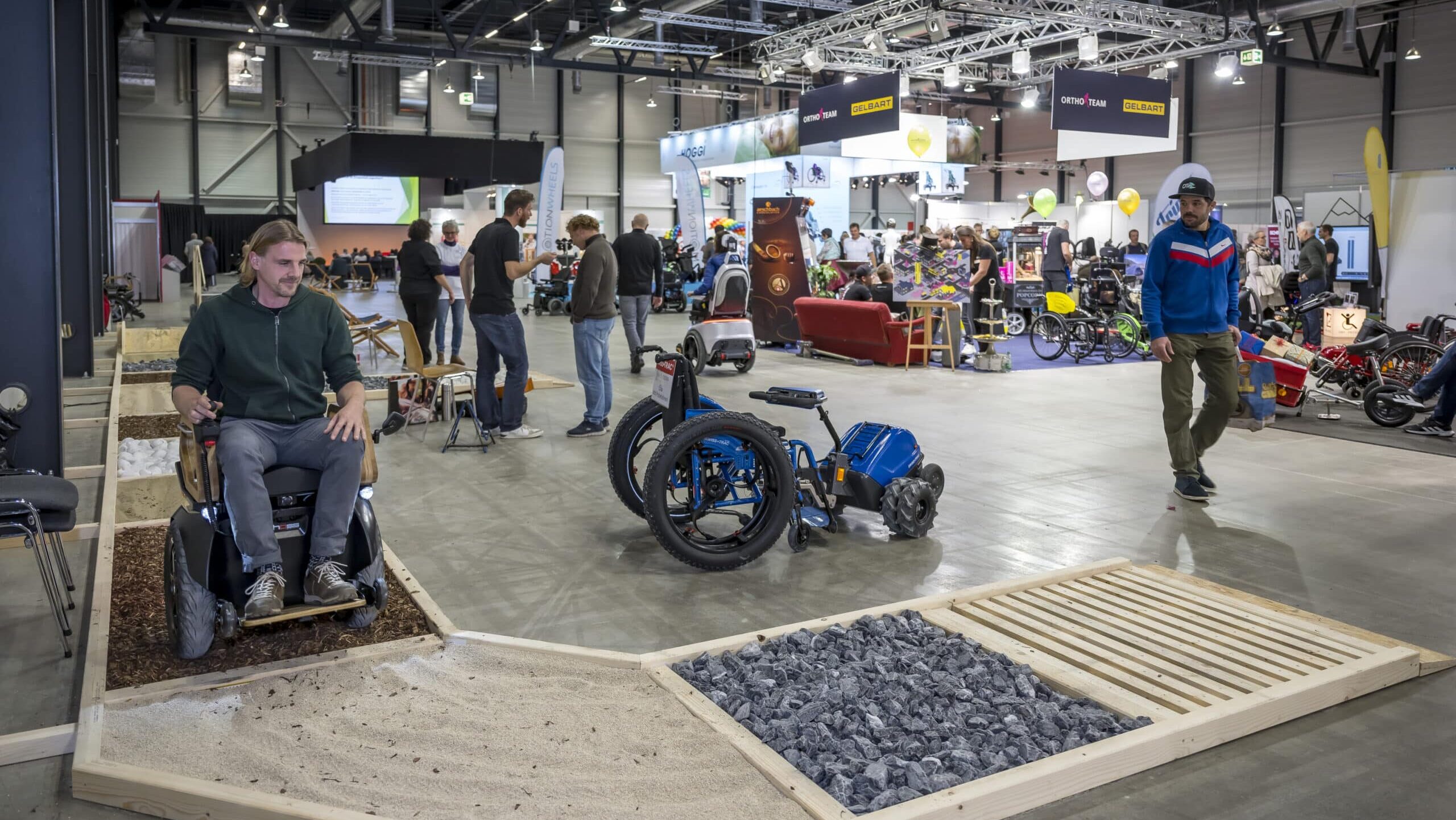 Ein Mobilitätsparcours an der Swiss Abilities. Weg mit verschiedenen Untergründen (Sand, Holzspäne, Steine), der in einer Messehalle ausgelegt ist. Auf dem Weg ein junger Mann im Rollstuhl. Im Hintergrund sieht man weitere Messestände.