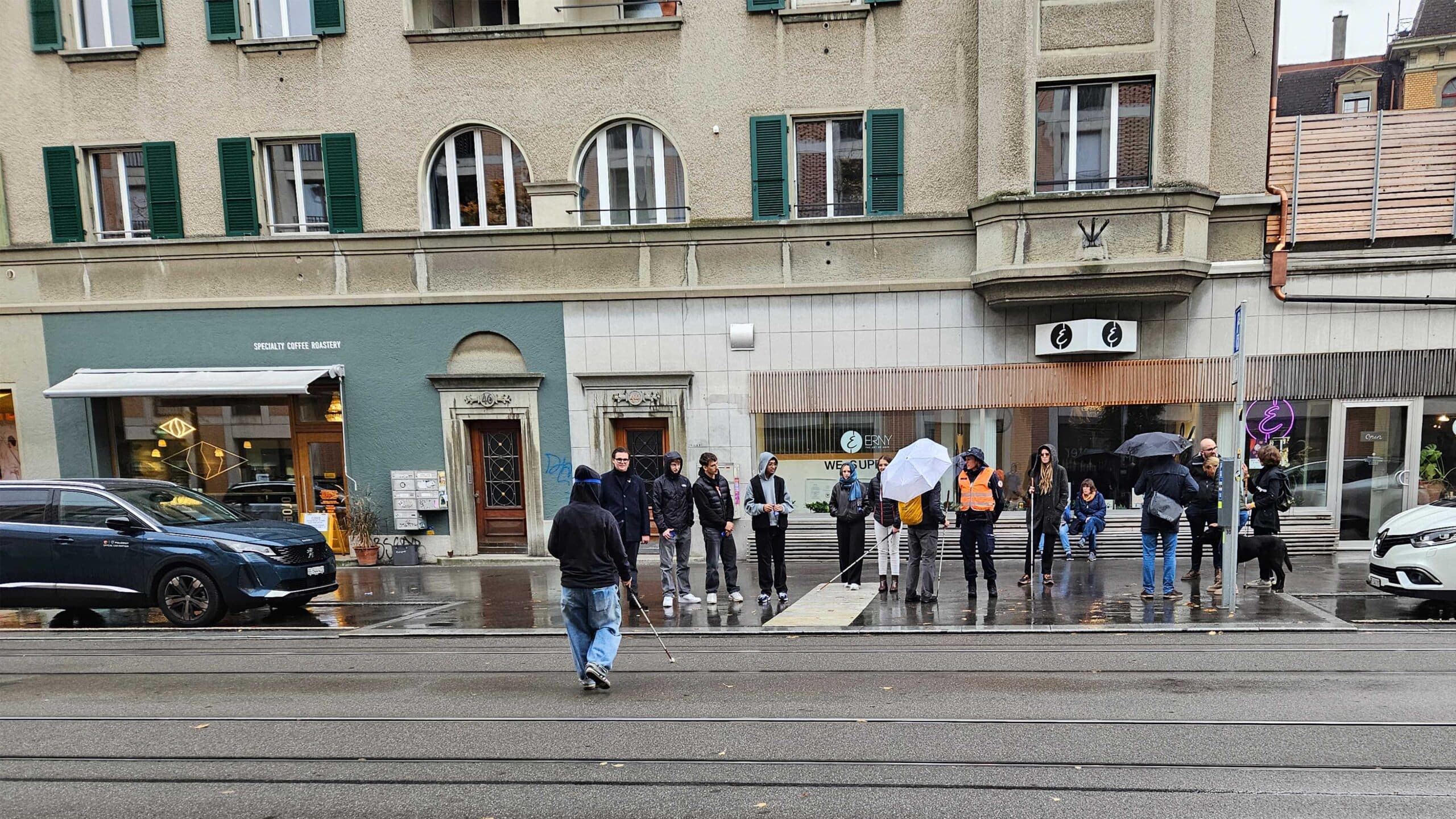 Eine Gruppe Jugendlicher steht am Strassenrand und sieht zu, wie eine sehbehinderte Person die Strasse überquert. 