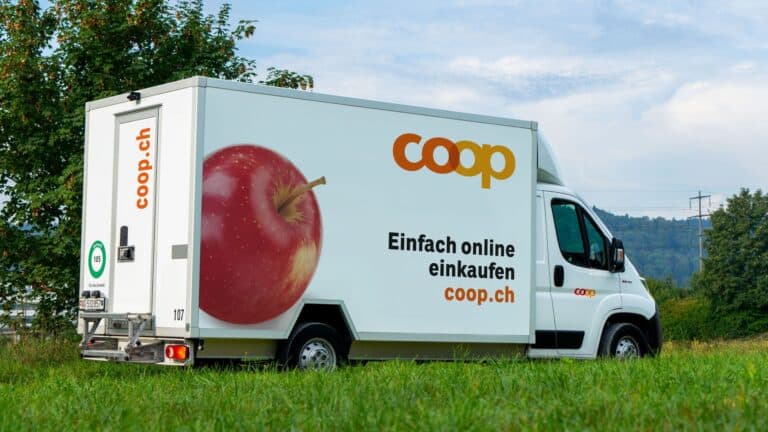 Ein weisser Coop-Lieferwagen fährt durch die Landschaft. Auf dem Lieferwagen steht: "Coop; Einfach online einkaufen; coop.ch". Links daneben ist ein grosser roter Apfel aufgedruckt.