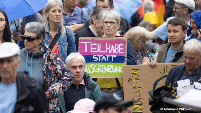 Menschenmenge, eine Person hält ein Schild hoch: "Teilhabe statt Ausgrenzung"