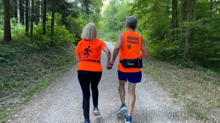 Eine Frau ist mit einem Guide am Joggen im Wald. Sie tragen beide orange Shirts. Man sieht die beiden nur von hinten. 