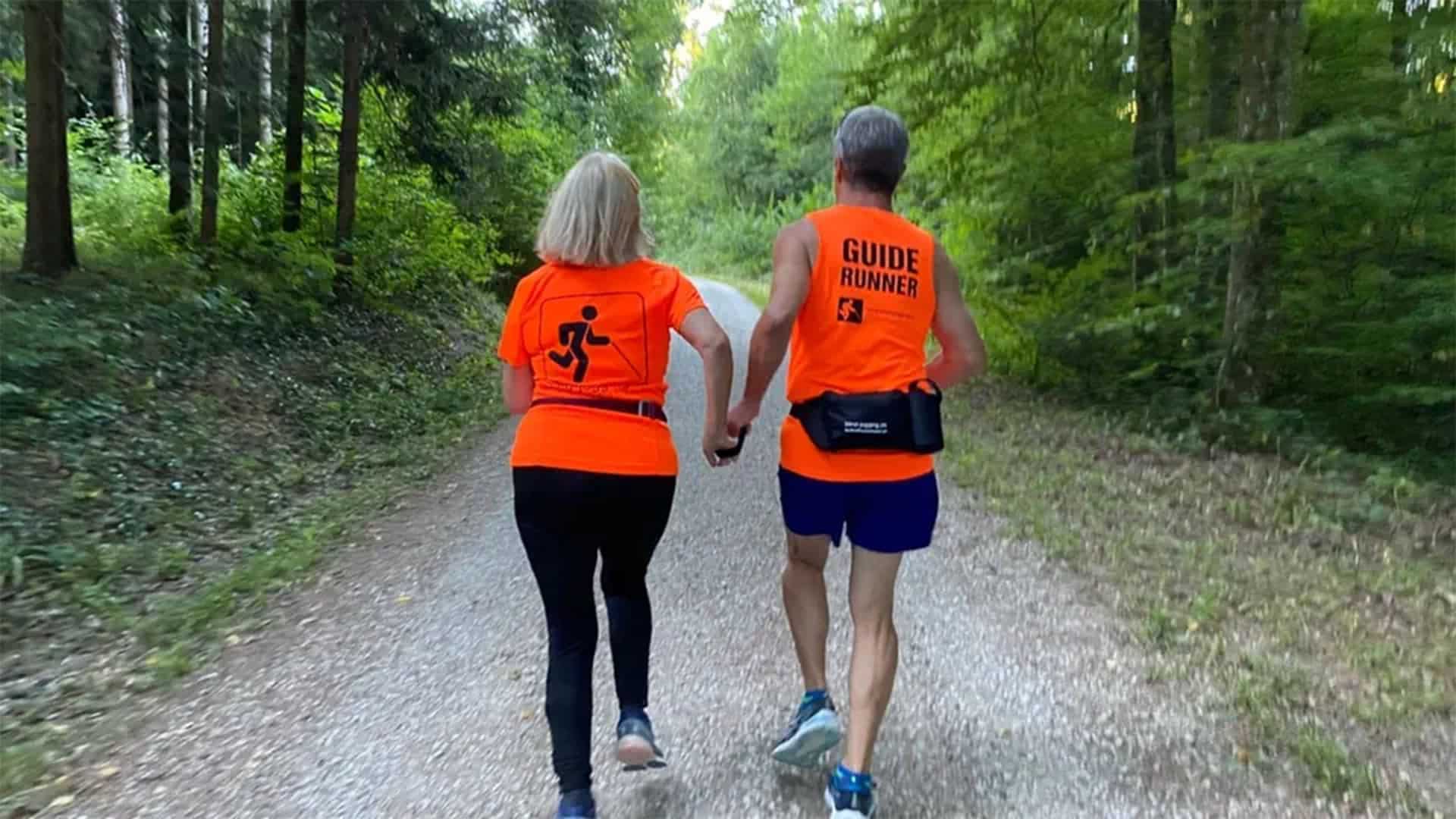 Eine Frau ist mit einem Guide am Joggen im Wald. Sie tragen beide orange Shirts. Man sieht die beiden nur von hinten. 