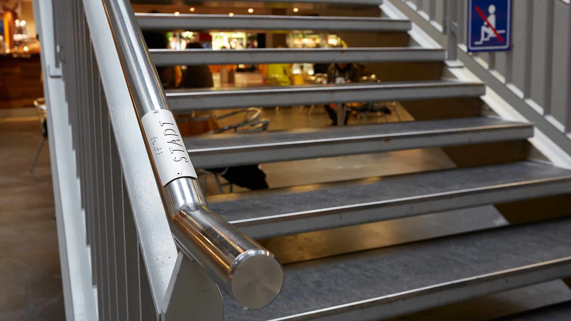 L'image montre un escalier dans la gare de Berne. Sur la rampe de l'escalier, on peut voir une inscription sur la main courante qui marque le chemin vers la ville.