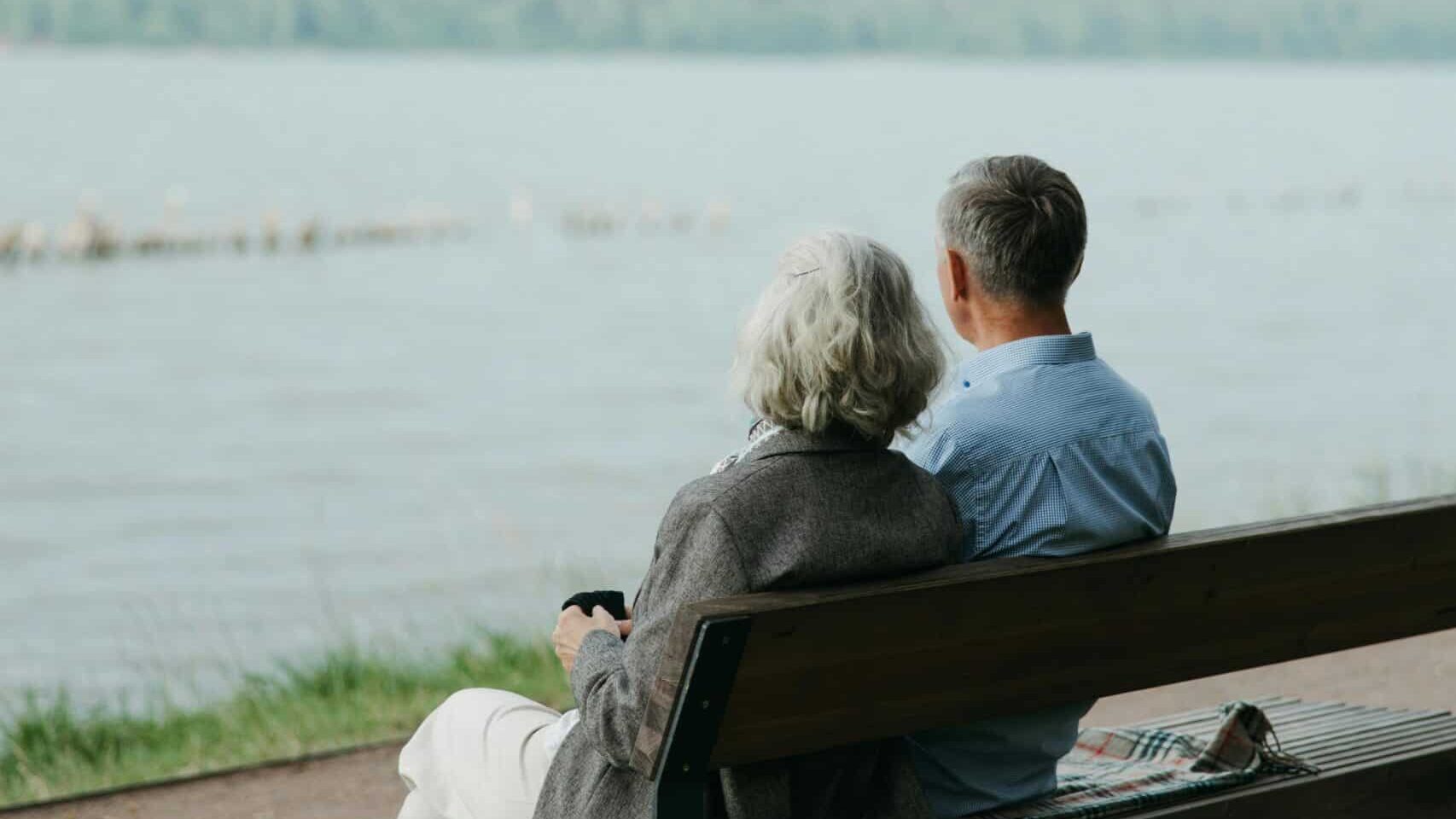 Ein älteres Paar sitzt auf einer Bank an einem See und schaut in die Weite. 