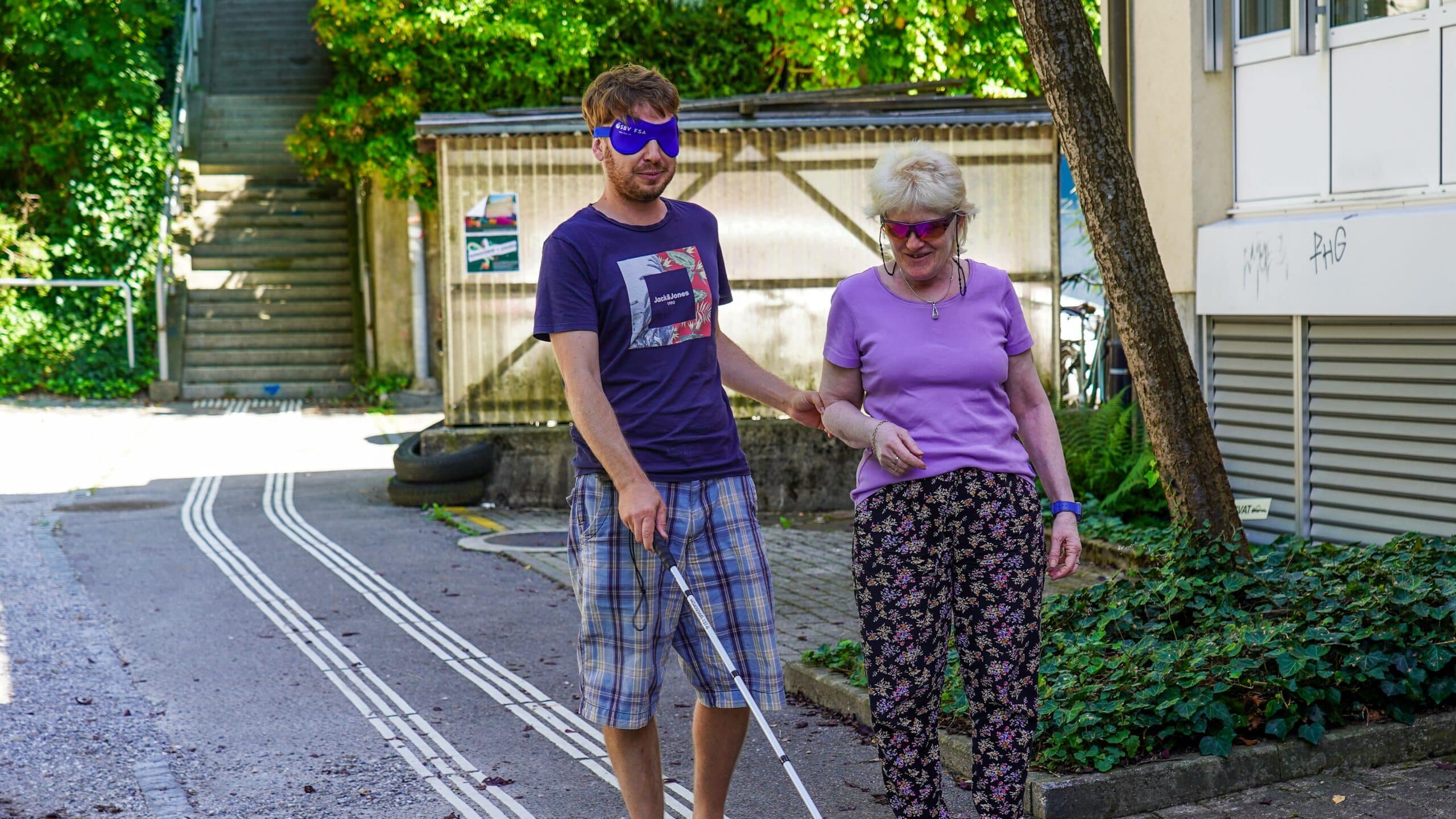 Mann mit Dunkelbrille und weissem Stock wird von einer sehbehinderten Frau geführt