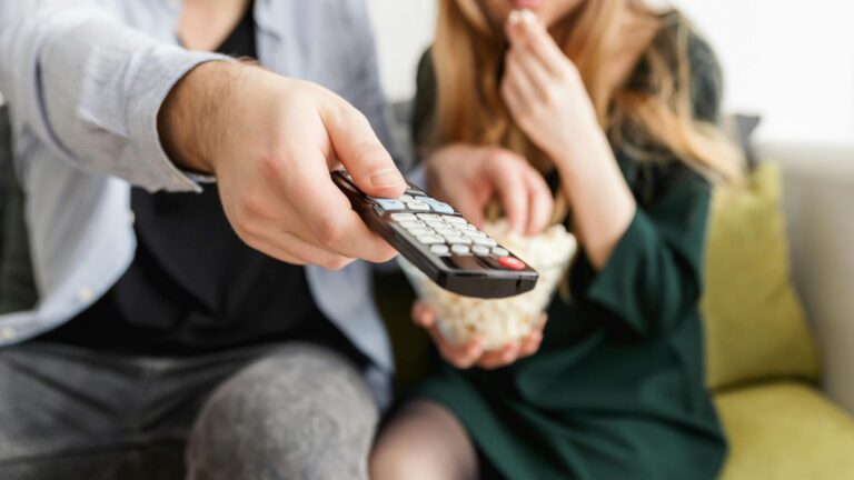 Zwei Personen sitzen mit Popcorn auf dem Sofa, man sieht nur ihre Körper, der Mann druckt die Fernbedienung aus