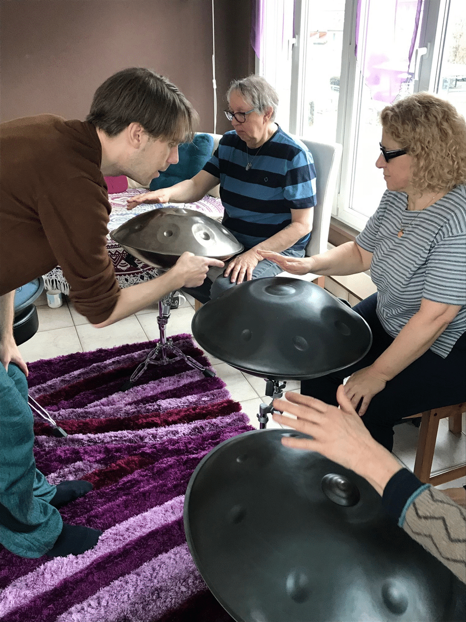 Deux participantes sont assises devant un handpan chacune et tapotent doucement l’instrument d’une main, tandis que le formateur semble expliquer quelque chose à l’une d’elles en lui montrant un geste de la main.