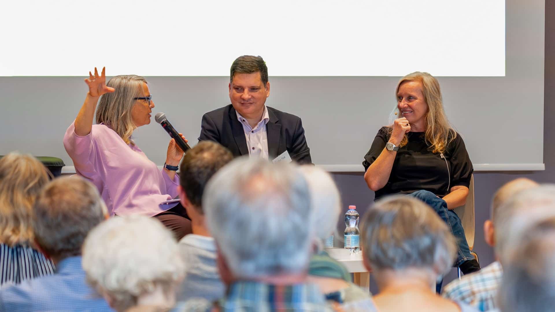 Blick durchs Publikum auf die Bühne. Drei Personen sitzen dort und diskutieren miteinander.