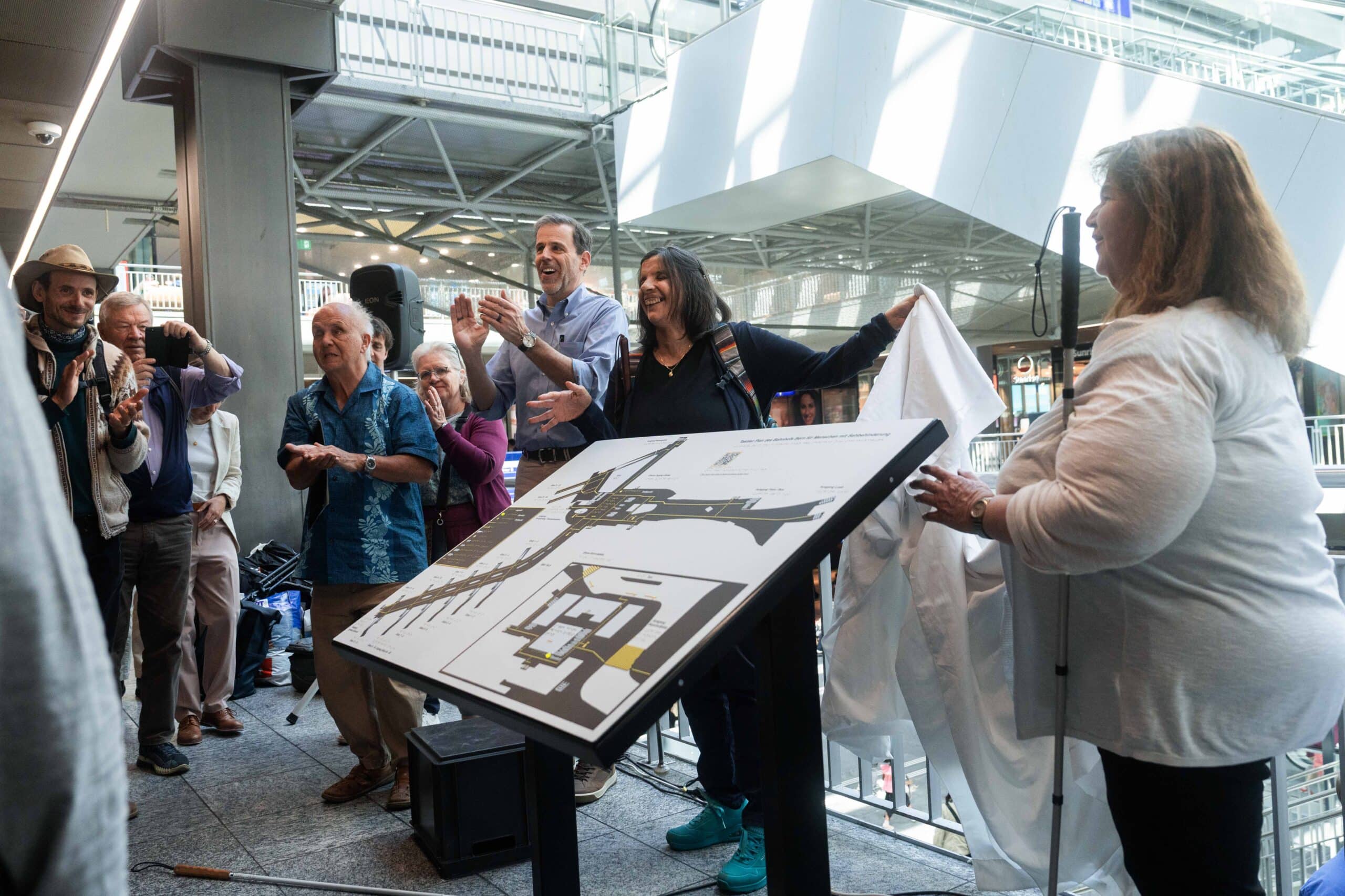 Moment, in dem das taktile Modell enthüllt ist: Frau hält Leintuch hoch, das das Modell bedeckt hatte, darum herum stehen Leute und applaudieren, es ist eine graue Platte ca. 1 Meter mal 70 cm mit Hervorhebungen und Braille-Beschriftung