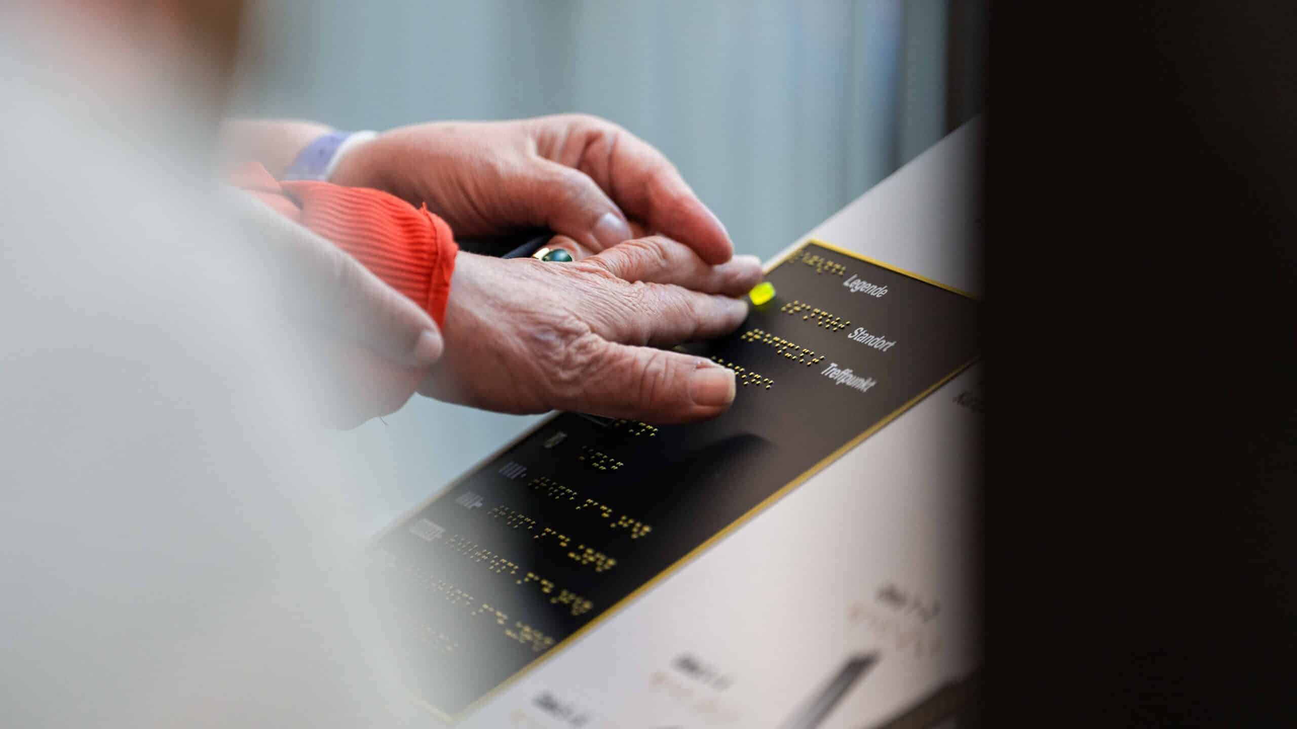 Zwei Hände auf einem Schild mit Braille-Schrift: Die obere Hand hilft der unteren über die Schrift zu gleiten.