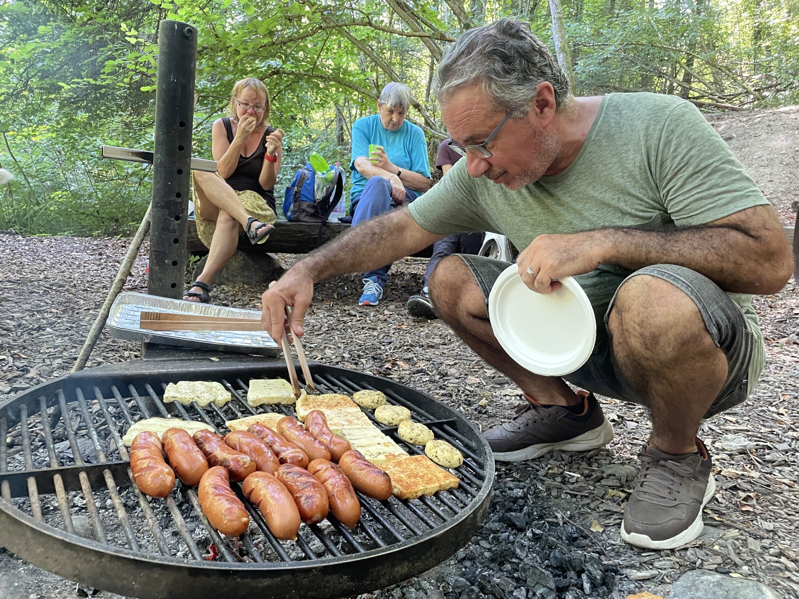 FOTO: Auf dem grossen, runden Grillrost der öffentlichen Feuerstelle liegen knusprige Cervelat-Würste sowie vegetarisches Grillgut bereit. Yusef in kurzen Hosen sorgt mit der Grillzange dafür, dass nichts anbrennt. Im Hintergrund sitzen zwei Frauen auf der Holzbank und warten hungrig auf das Essen.