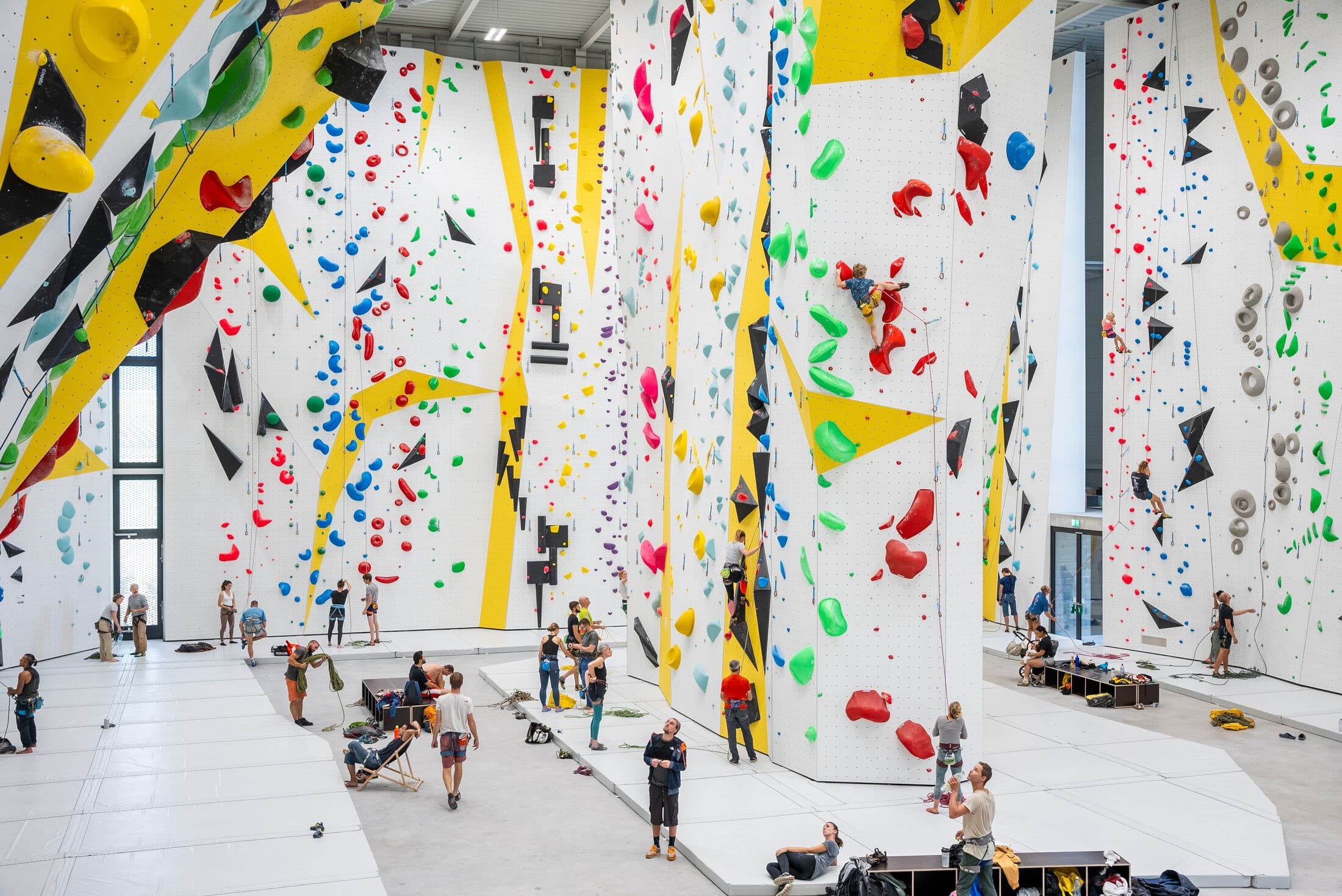 Des personnes dans une grande salle d’escalade intérieure, grimpant sur des murs colorés avec prises variées; ambiance sportive et dynamique.
