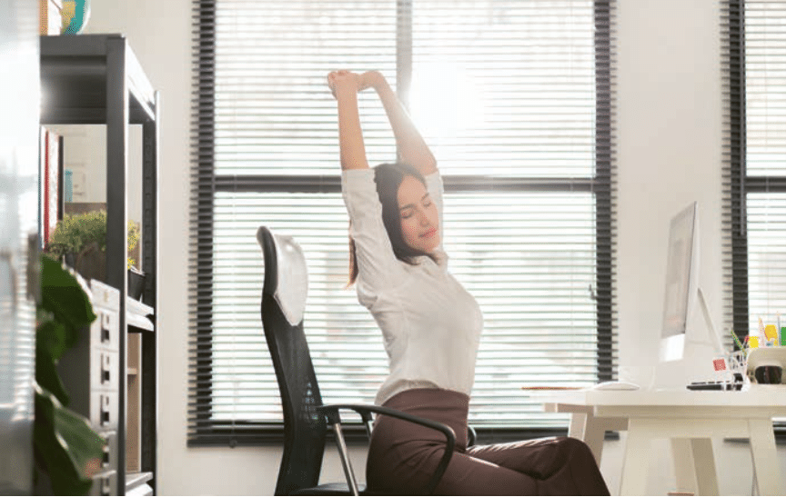 Une femme assise à son bureau s’étire avec les bras levés, les yeux fermés, profitant d’un moment de détente dans un bureau lumineux avec de grandes fenêtres.