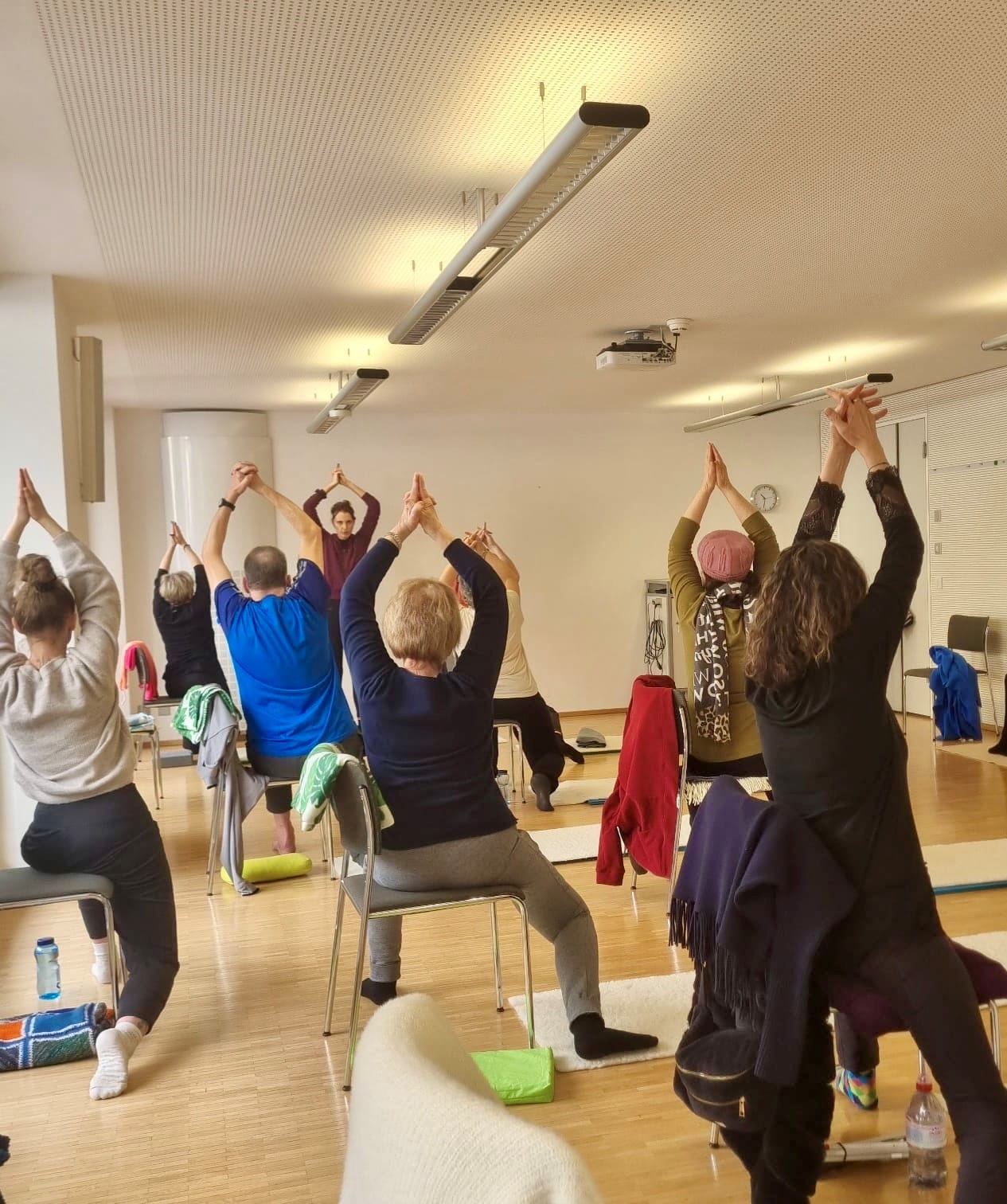 Un groupe pratique un exercice assis dans une salle lumineuse à Interlaken. À l’avant, la personne qui dirige le cours montre la posture, tandis que les participants suivent le mouvement.