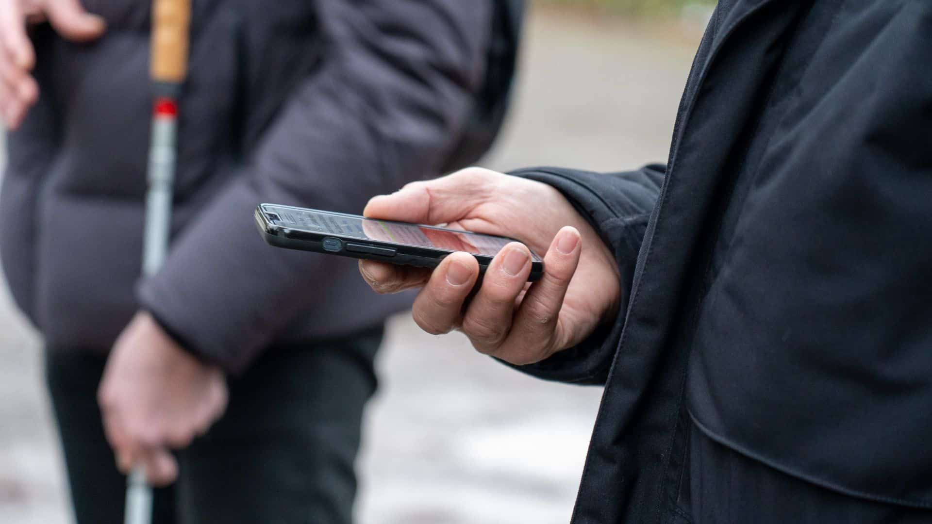 Im Mittelpunkt des Bildes ist eine Hand die ein Mobiltelefon hält. Im Hintergrund ist jemand mit einem weissen Stock.