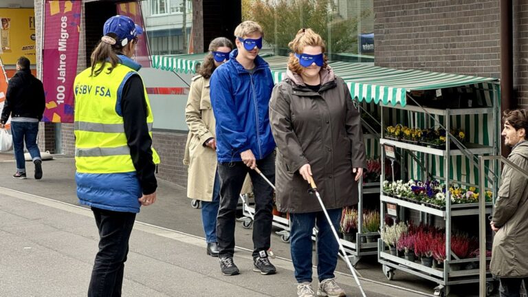 Rechts laufen 3 Personen mit weissem Stock und Dunkelbrille hintereinander. Links steht eine Frau mit gelber Weste mit Aufdruck sbv fsa und leitet.