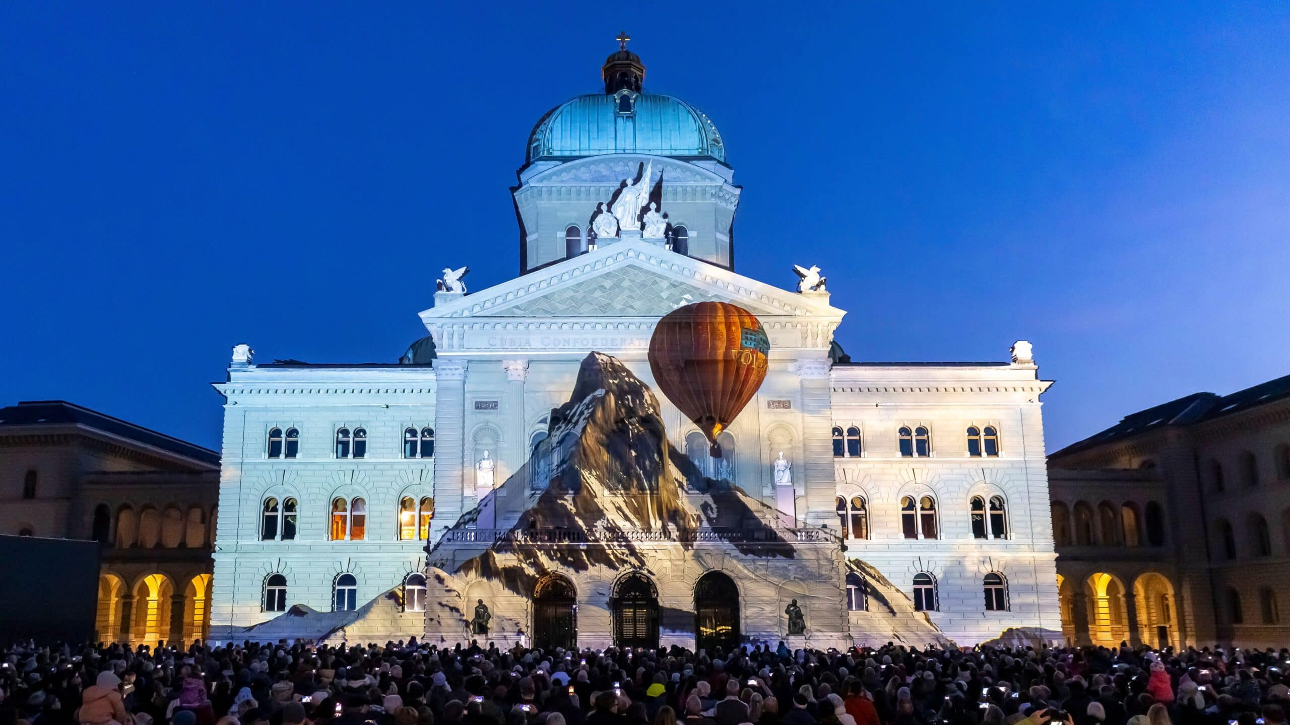 Photo prise lors du spectacle lumineux. Le Palais fédéral est illuminé en blanc dans un ciel crépusculaire sans nuages, avec au centre le Mattherhorn, au-dessus duquel flotte une montgolfière.   