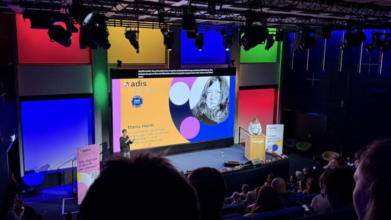 Blick aus dem Publikum auf die Bühne der Tagung; Leinwand und Rednerpult sind im Hintergdun von bunten Leuchtkacheln in den Google-Farben gerahmt.