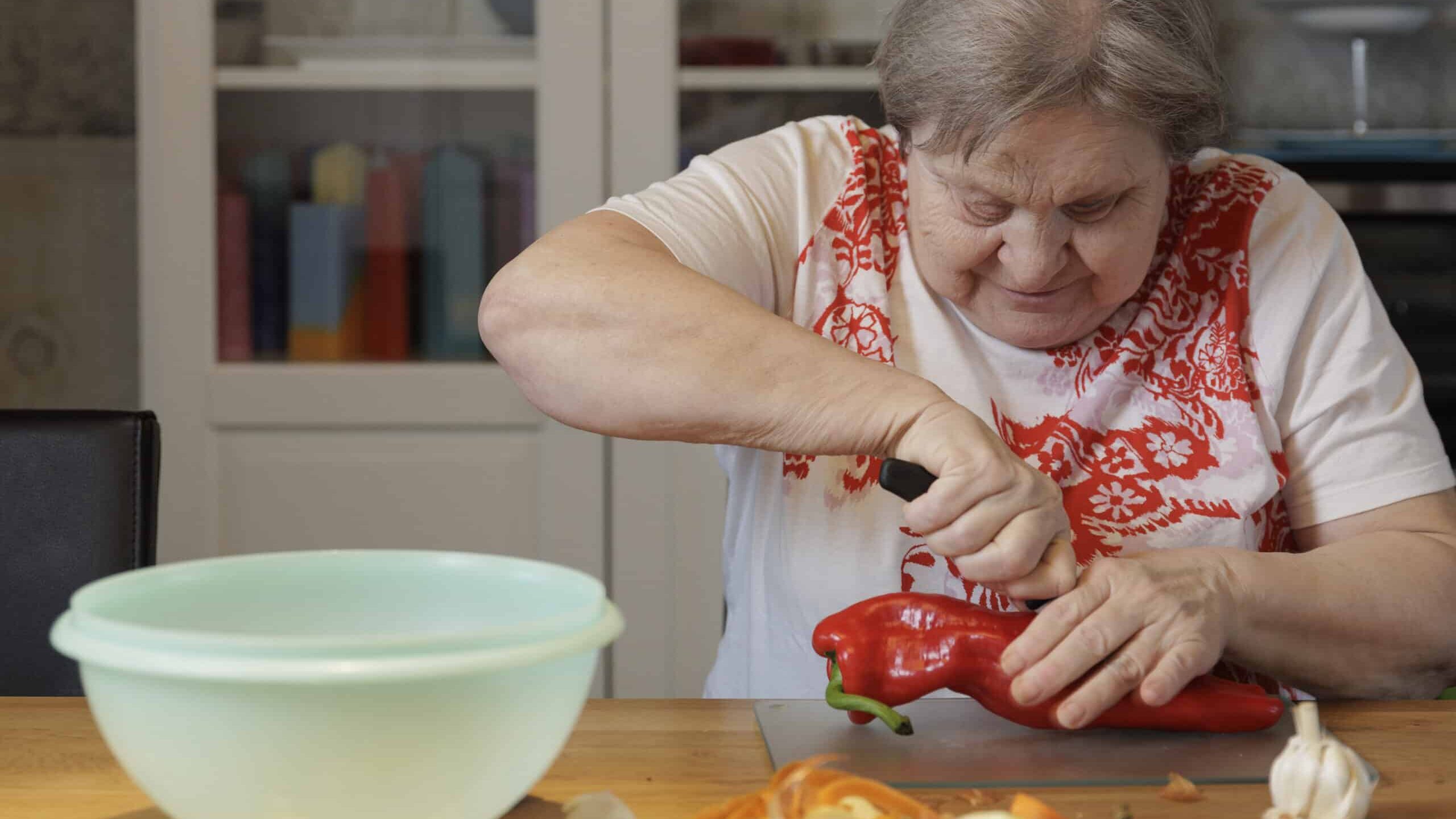 Jolanda Gehri sitzt in einem weissen T-Shirt mit roten Blumenmustern an ihrem Küchentisch. Konzentriert schneidet sie eine riesige rote Paprika der Länge nach auf und legt sie zu den bereits geschnittenen Knoblauchzehen, Zwiebeln und gelben Paprikaschoten.