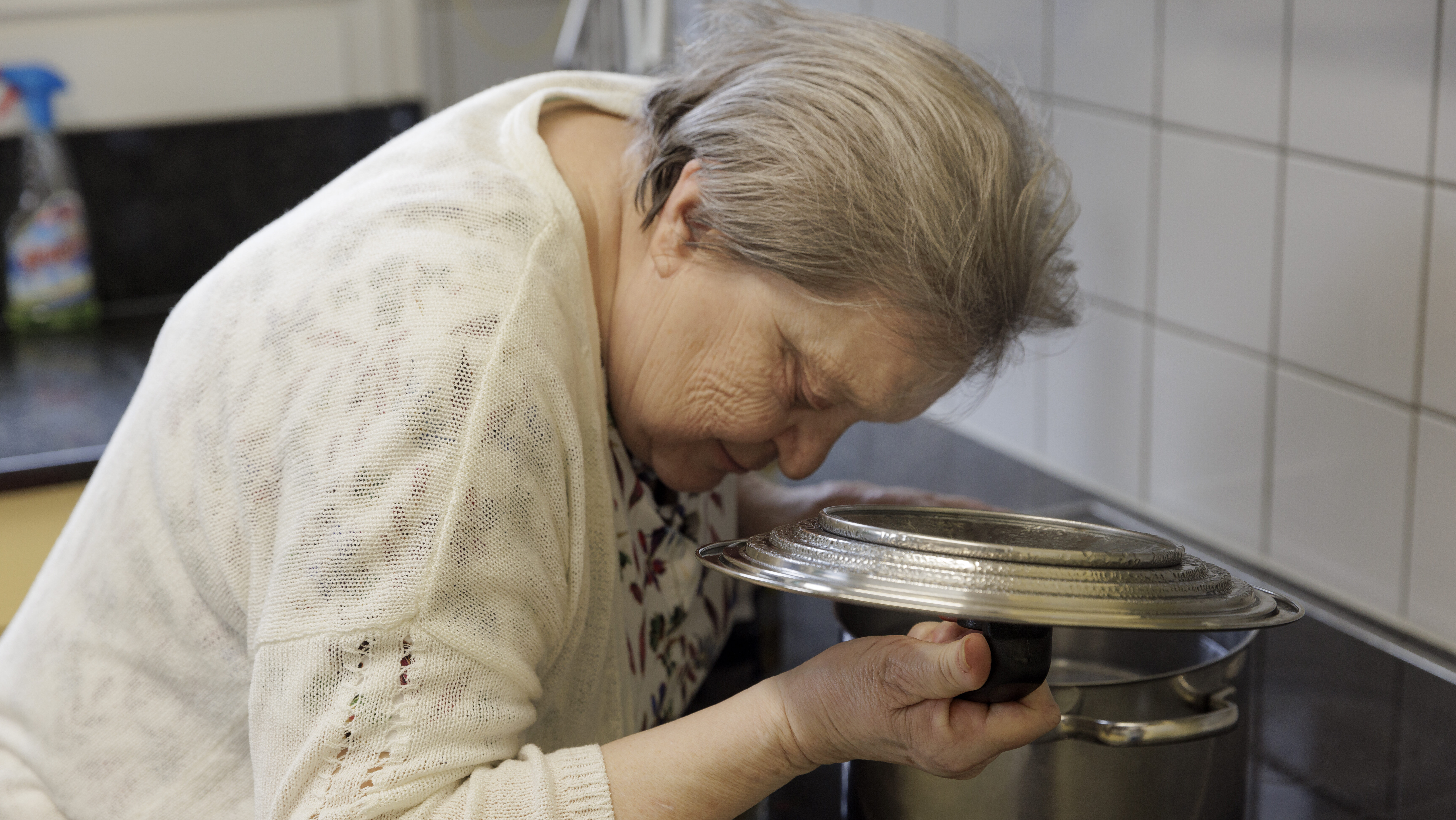 Eine ältere Dame beugt sich über ihren Herd und kontrolliert lauschend, wie der Inhalt eines grossen Topfes kocht: Sie sich vor dem Dampf, indem sie einen Deckel über den Topf hält.