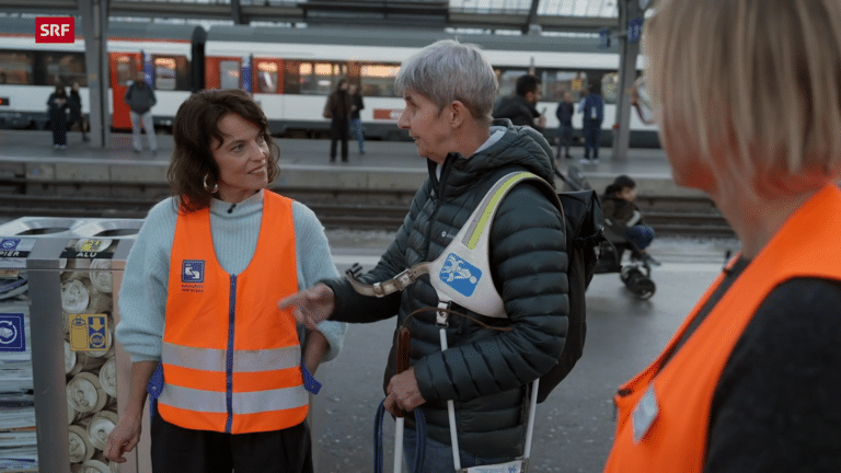 TV-Beitrags-Ausschnitt: Moderatorin Mona Vetsch (links, mit orangener Leuchtweste) spricht mit Domenica Griesser (rechts, trägt Gstältli vom Führhund über der Schulter). Im Hintergrund der Bahnhof Zürich mit Perrons und einem Zug.