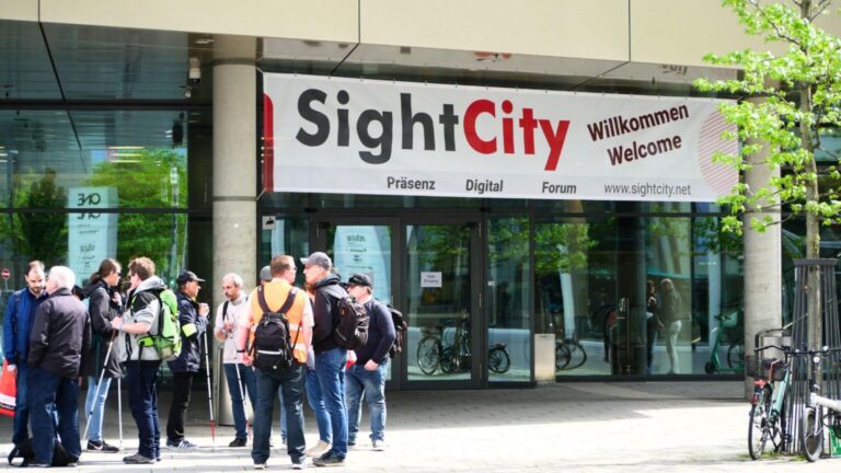 Un groupe de personnes, dont beaucoup portent une canne blanche, se tient devant une façade en verre avec une porte d'entrée surmontée d'une bannière "SightCity - Willkommen -Welcome" (en plus petit : präsenz digital forum www.sightcity.net).