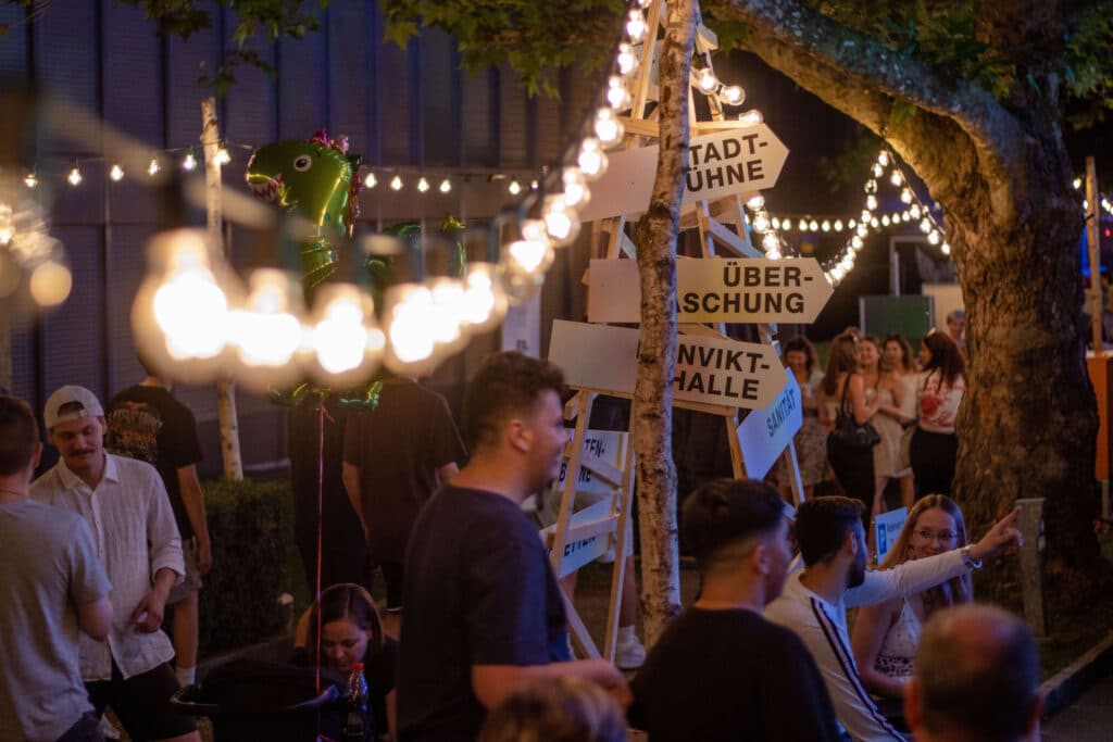 Impression des Fests aus dem Vorjahr, man sieht Menschen, abends, an Ständen und Bars, eine Lichterkette und im HIntergrund Holz-Wegweiser 