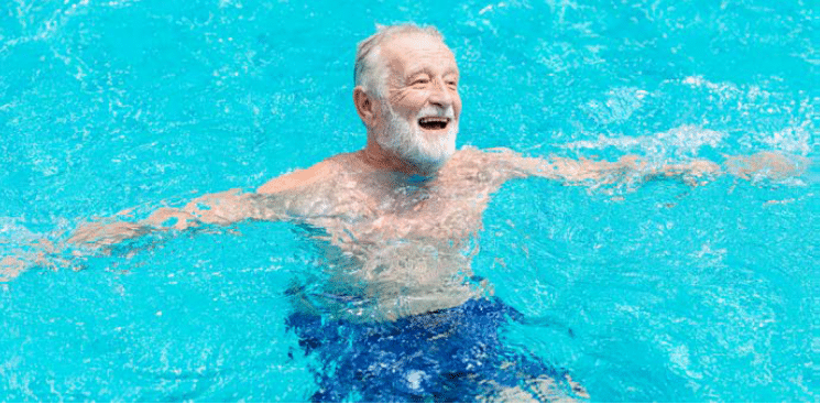Un senior aux cheveux blancs et barbu dans l'eau d'une piscine, qui a l'air de rire aux éclats.