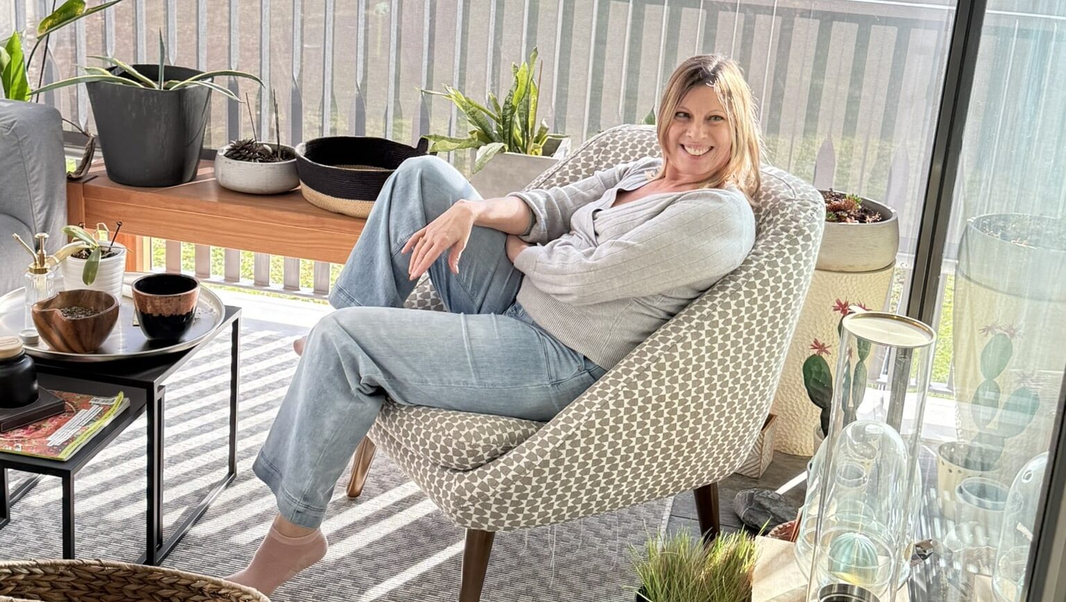 In einer sonnendurchfluteten Loggia, umgeben von Pflanzen, sitzt eine blonde Frau lächelnd und entspannt in einem gepolsterten Sessel.