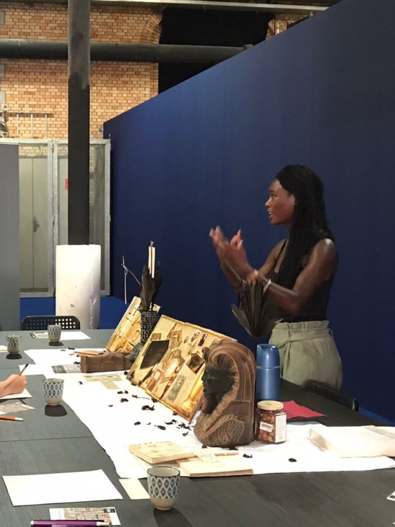 Une femme, historienne de l’art et formatrice, explique avec enthousiasme les images historiques d’Égypte qui se trouvent devant elle. Elle se tient devant une longue table noire, derrière elle une une paroi bleue est installée. Sur la table, on distingue également des sculptures emblématiques de l’Égypte, qui complètent l’illustration des images.