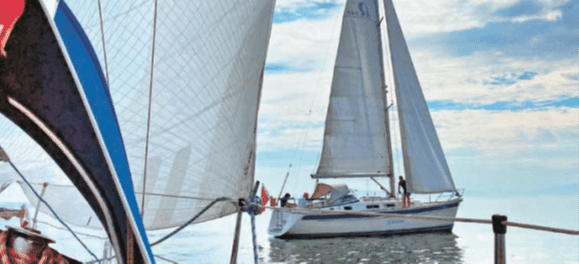 Deux voiliers naviguent sur une eau calme sous un ciel partiellement nuageux, l’un vu de près et l’autre avançant au large.