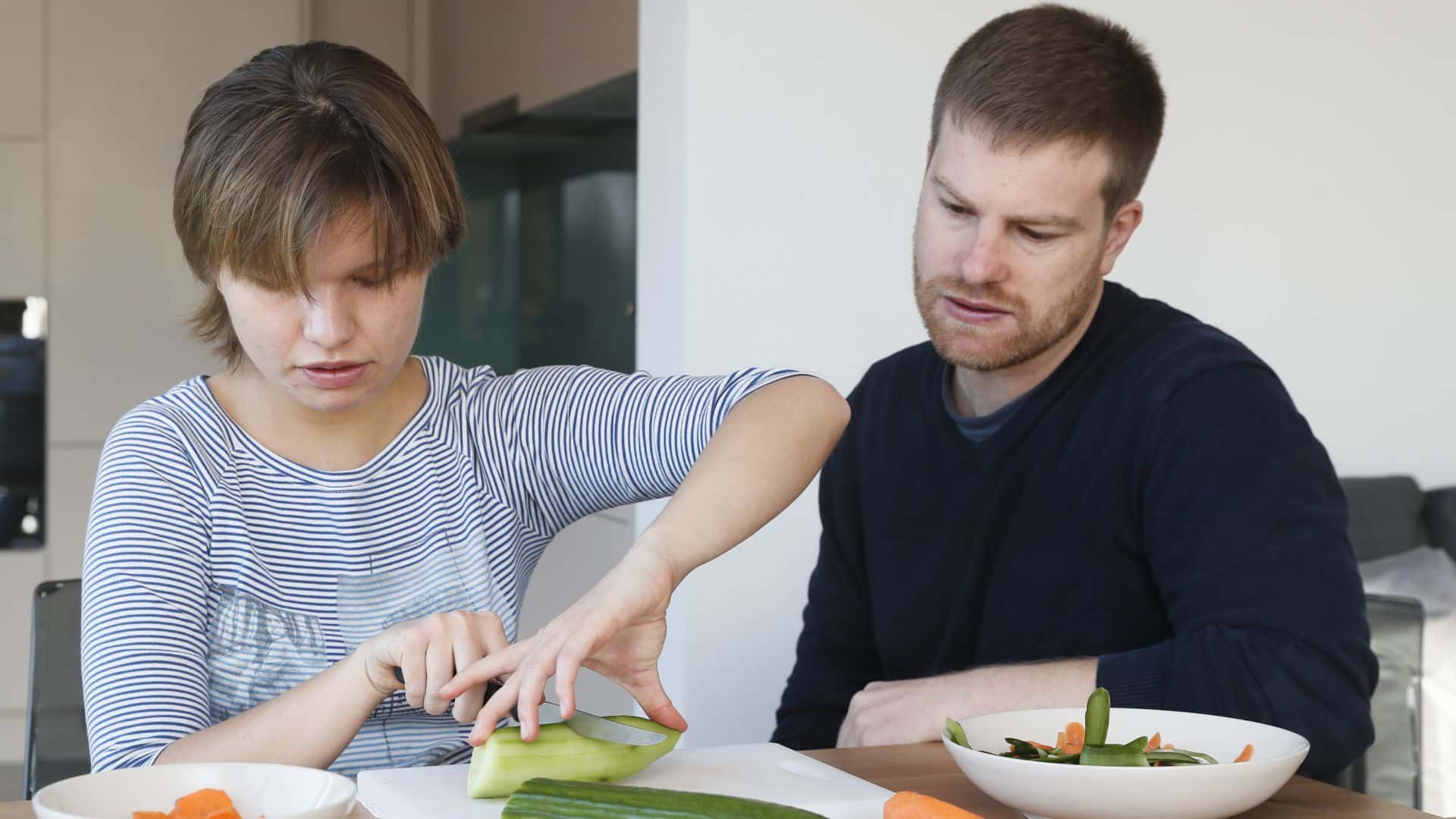 In einer gut beleuchteten Küche schneidet eine junge blinde Frau mit geneigtem Kopf Gemüse unter dem aufmerksamen Blick ihres LPF-Beraters.
