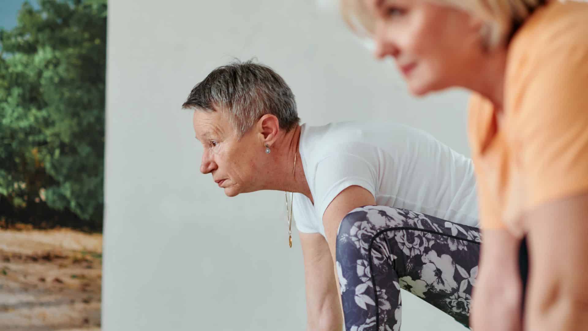 Zwei Personen üben eine stabile Yoga-Position. Die sehbehinderte Person im Fokus beugt sich kontrolliert und mit fokussiertem Ausdruck nach vorne und stützt die Hände auf der Matte.