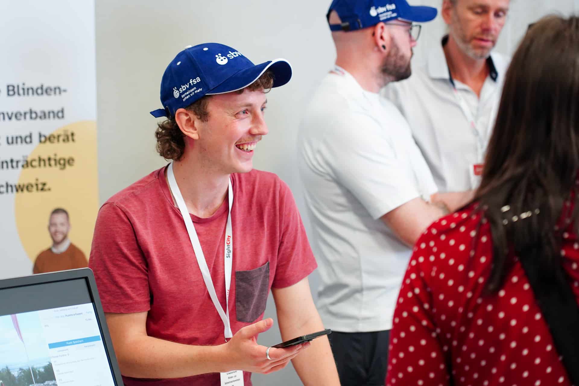 Stand des sbv. Eine Person mit blauem sbv-Cap erklärt lacht und erklärt einer Frau eine App am Smartphone.
