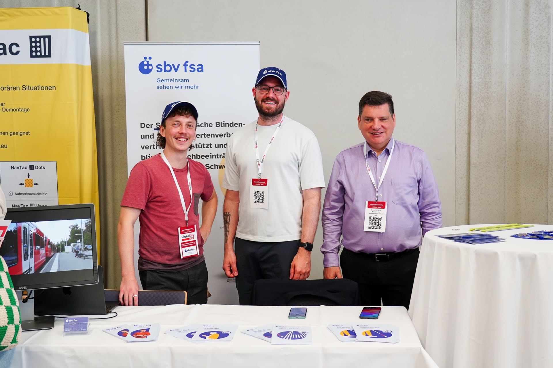 Stand des sbv an der SightCity 2026. 3 Männer aus der T&I-Abteilung stehen lachend hinter einem weissen Tisch. Auf dem Tisch liegen Flyer und link läuft auf einem Bildschirm ein Video
