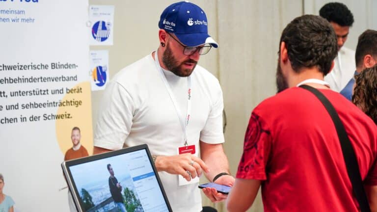 Stand des sbv. Eine Person mit blauem sbv-Cap erklärt einem interessierten Mann die App und tippt auf das Smartphone.
