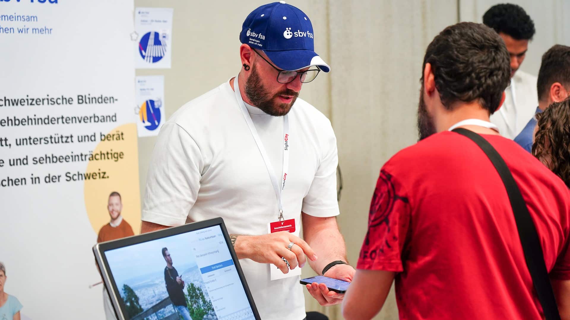 Stand des sbv. Eine Person mit blauem sbv-Cap erklärt einem interessierten Mann die App und tippt auf das Smartphone.
